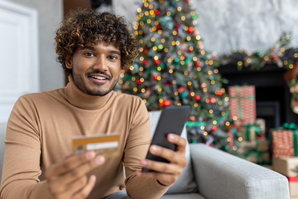 Portrait of happy satisfied Christmas shopper, Hispanic man smiling and looking at camera sitting on sofa in living room at home, man holding bank credit card, using smartphone app for shopping